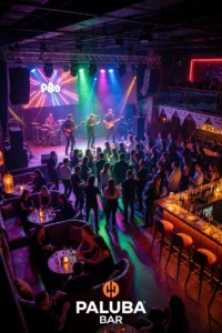 A vibrant nightclub scene at Paluba Bar, featuring a lively crowd dancing under colorful neon lights, with a prominent stage area for live performances, and a separate lounge section with comfortable seating and a bar, all set on October 31st with Halloween-themed decorations